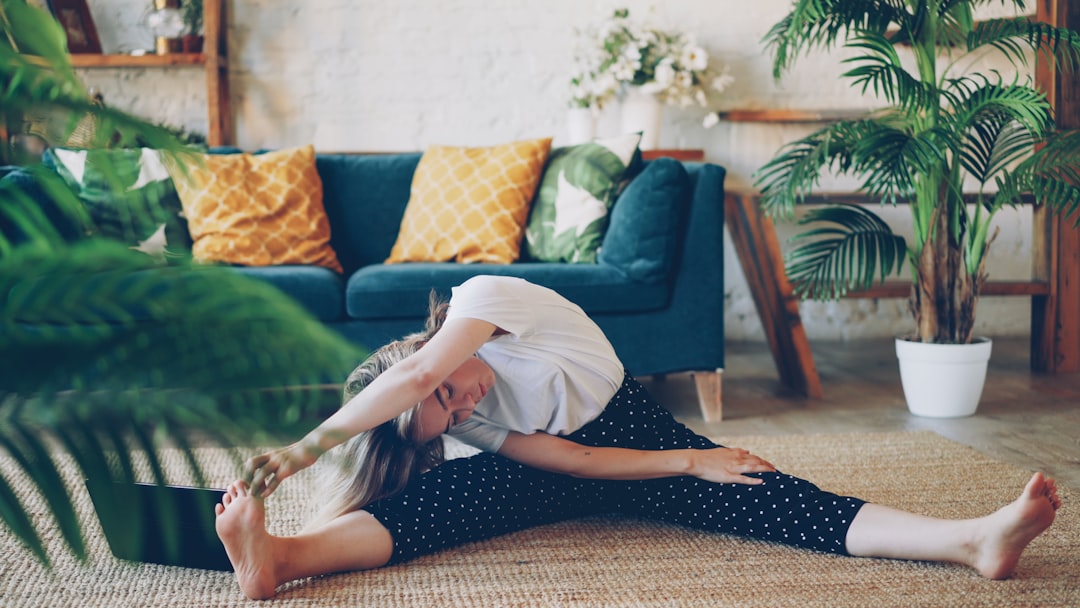 facilities Flexible young blonde is doing stretching exercises at home sitting on floor in modern apartment. Girl is bending head to knee holding toes and stretching back and legs.